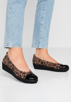 Fødder iført kilehælssko med leopardmønster og skinnende sorte tåkapper, parret med lyseblå cropped jeans, stående på et hvidt gulv.