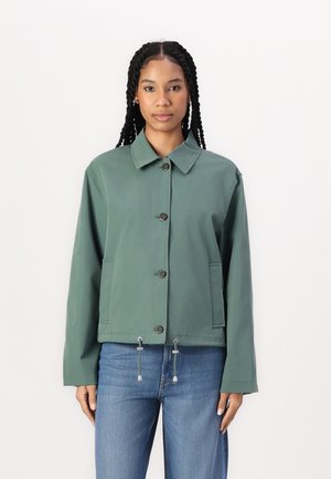 Grønn kort jakke med knapper foran, spiss krage, sidelommer og snorjusteringer i kanten, parret med blå jeans.