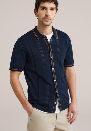 Homme portant une chemise à manches courtes bleu marine avec bordure marron sur un sous-vêtement blanc et un pantalon beige, debout avec une main dans la poche.