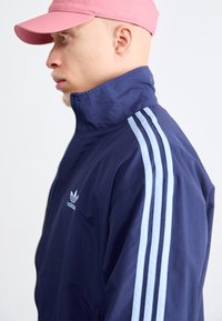Tamsiai mėlynas vėjyje atsparus striukė su aukštu kaklu, šviesiai mėlynas trijų juostų dizainas ant rankovių ir mažas baltas Adidas logotipas priekyje. Matoma rožinė kepurė.