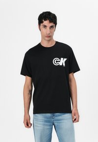 T-shirt gráfica de algodão preto com um logótipo branco "CK" no lado esquerdo do peito. Gola redonda, mangas curtas e ajuste descontraído. Usado com jeans azul claro.