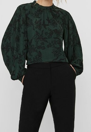 Grøn bluse med lange ærmer med sort blomstermønster og puffede ærmer kombineret med højtaljede sorte bukser, hænderne i lommerne.