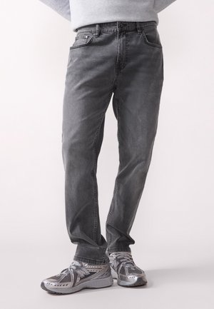 Man wearing grey washed straight-fit jeans and silver New Balance sneakers standing against a plain background.