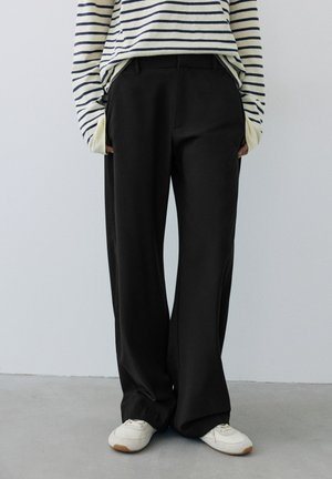 Personne portant un pantalon large noir, des baskets blanches et un haut blanc à manches longues avec des rayures horizontales noires, debout à l'intérieur.