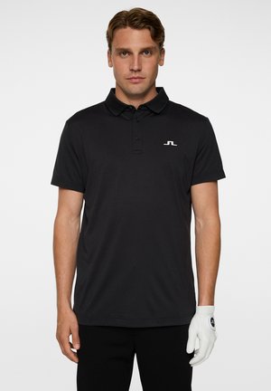 Sort polo-shirt med klassisk krave, kortærmede, og et diskret logo på brystet. Bæres med sorte bukser og en hvid golfhandske.