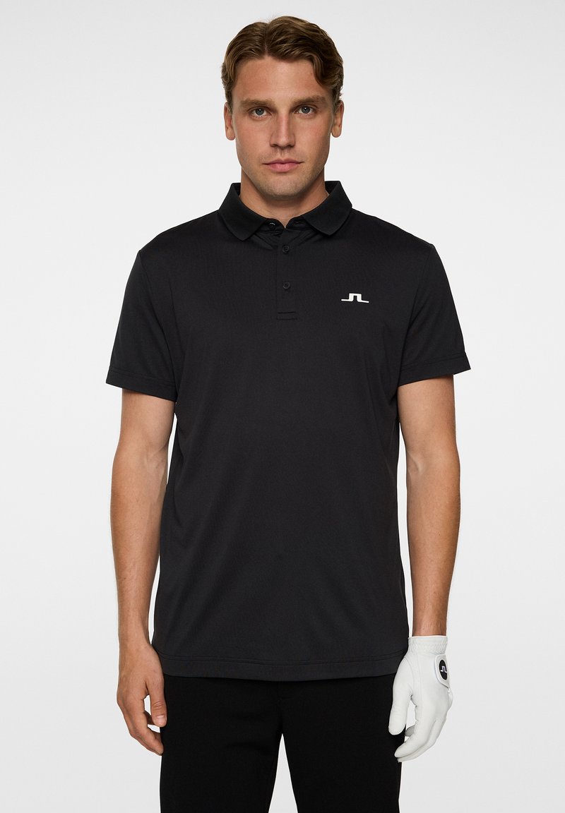 Musta polo-paita, jossa on klassinen kaulus, lyhyet hihat ja hillitty logo rinnassa. Yhdistetty mustiin housuihin ja valkoiseen golf-käsineeseen.