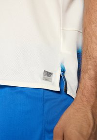 Witte sportshirt met een textuurpatroon, voorzien van een blauwe accent aan de zoom, naast een grijze New Balance logo patch aan de zijkant.