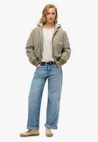Licht olijfgroen bomberjack met een grijze capuchon, gecombineerd met een licht shirt en losse blauwe jeans. Witte sneakers met beige accenten.