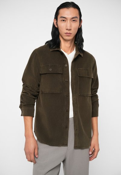 Camisa de cor de azeitona em veludo cotelê com dois bolsos frontais, fecho com botões e mangas arregaçadas, combinada com uma t-shirt bege por baixo.
