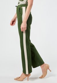 Green pants with a relaxed fit, featuring cream side stripes, elastic waistband, and pockets; worn with nude high-heeled shoes.