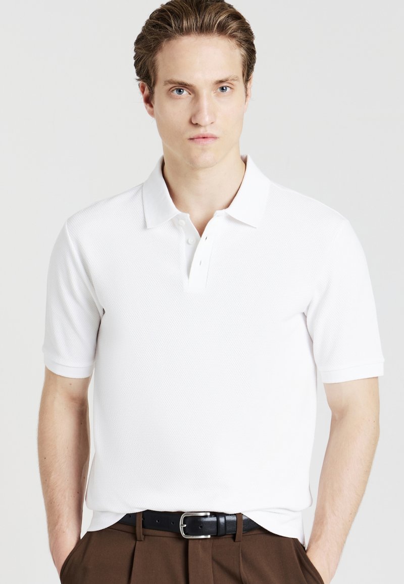 Polo bianco realizzato in tessuto strutturato con apertura a tre bottoni. Presenta maniche corte e colletto piatto. Indossato con pantaloni marroni e cintura nera.
