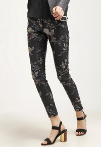 Mujer con pantalones negros ajustados con estampado floral y tacones abiertos negros con tacón grueso dorado, posando frente a un fondo liso.