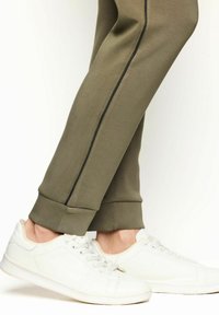 Oliwkowe spodnie dresowe o dopasowanym kroju, z czarnymi lampasami po bokach i ściągaczami na końcach, w połączeniu z białymi sneakersami.