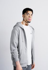 Sudadera con cremallera gris claro y bolsillos frontales, combinada con una camiseta blanca. Joven con pelo rizado corto posando frente a un fondo neutro.