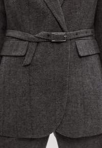 Sivi sako s herringbone uzorkom izbliza, koji ima prilagođeni dizajn, oštre reveri, pojas s srebrnom kopčom i bočne džepove.