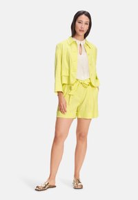 Lime green linen blazer with three-quarter sleeves, paired with matching shorts featuring a tie waist, and a light beige top. Brown patterned sandals.