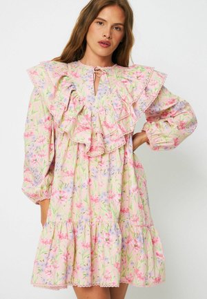 Ženska, oblečena v pastelno cvetlično obleko z večplastnimi volani, dolgimi napihnjenimi rokavi in majhnim izrezom v obliki ključavnice s trakom.