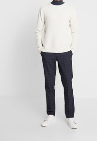 Un pull crème avec un design en tricot texturé, associé à un pantalon à carreaux navy et des baskets blanches, affichant un look décontracté mais soigné.