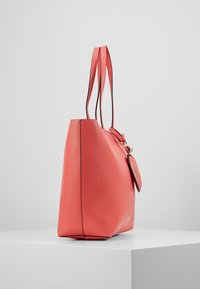 Sac fourre-tout corail avec une finition texturée, une base plate et deux bandoulières. Dispose de ferrures dorées et d'une étiquette amovible en accent.