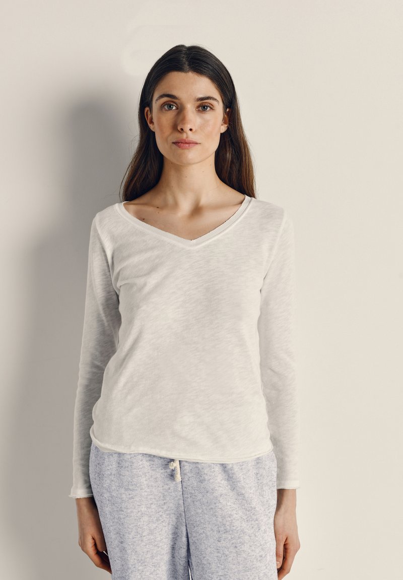 Mlada žena s dugom tamnom kosom, odjevena u bijelu majicu dugih rukava s V-izrezom i svijetlosive trenirke, stoji nasuprot jednobojnom bež zidu.