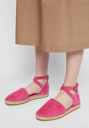 Roze suède espadrille platte schoenen met enkelriempjes en gesp, gedragen met een beige rok, getoond op lichtgekleurde benen tegen een grijze achtergrond.