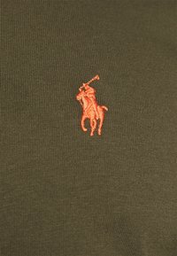 Tkanina w oliwkowej zieleni z wyraźnym czerwonym haftowanym logo polo gracza, prezentująca teksturowany, prążkowany wzór.