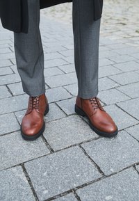 Smeđe kožne gležnjače s vezanjem, okruglim vrhom i crnom gumenom potplatom, uparene s ljubičastim krojenim hlačama na kamenom površinu.