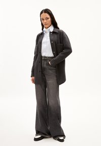 Camicia giacca oversize in misto lana grigia con tasca, indossata sopra una camicia bianca a maniche lunghe. Abbinata a jeans flare grigio scuro a vita alta e scarpe nere.