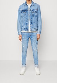 Chaqueta de mezclilla azul claro con cierre de botones y dos bolsillos en el pecho, combinada con jeans azules claros desgastados y zapatillas blancas.