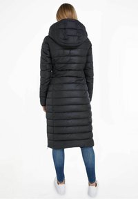 Black puffer coat with a hood, featuring a quilted design and knee-length cut. Worn with blue jeans and white sneakers.