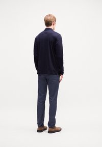 Marineblaues Langarmhemd aus weichem Stoff, gerade geschnitten. Kombiniert mit grauen Hosen und braunen Schuhen, von hinten vor einem hellen Hintergrund betrachtet.