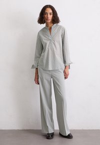 Blouse à manches longues avec un motif circulaire vert et blanc, présentant un col mandarin et des poignets volants. Associée à un pantalon large gris clair.