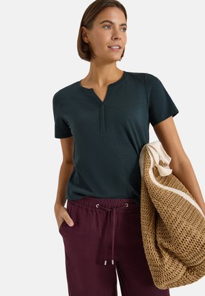 Femme portant un t-shirt bleu sarcelle foncé à manches courtes et un pantalon bordeaux à cordon, tenant un grand sac cabas tissé beige sur le bras.