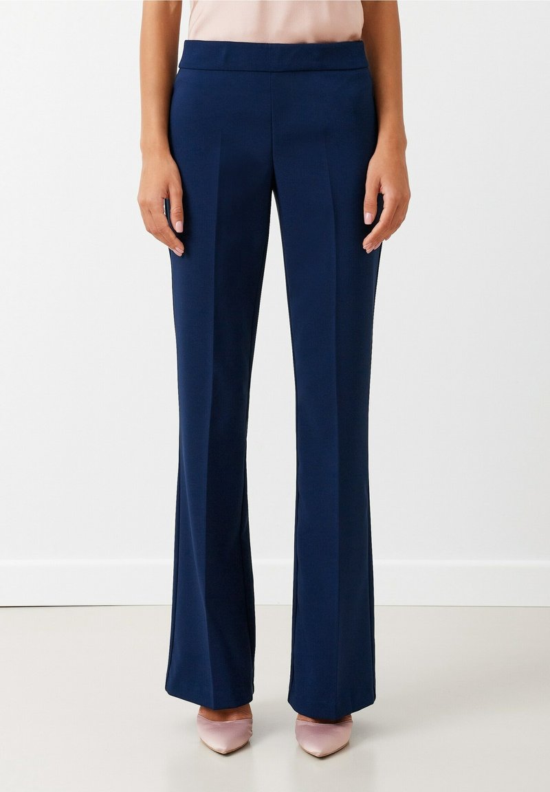 Femme portant un pantalon large sur mesure bleu marine et des escarpins rose pâle à bout pointu, debout contre un mur blanc uni.