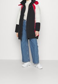 Person iført en sort, rød og hvid farveblokeret vinterfrakke, hvid rullekrave, blå vide jeans og hvide sneakers på ensfarvet baggrund.