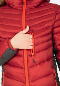 Rød isoleret jakke med horisontale syninger, kontrasterende orange lynlås og grå ærmer. Har en lynlåslomme på brystet.