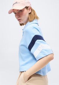 Νεαρή γυναίκα με ανοιχτό μπλε κοντομάνικο πουκάμισο με ναυτικές και λευκές ρίγες, μπεζ παντελόνι και ροζ καπέλο, στέκεται με το χέρι στην τσέπη της.