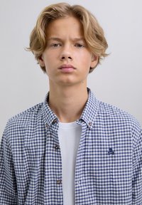 Joven rubio con cabello de longitud media, vistiendo una camisa de cuadros azul y blanca sin abotonar sobre una camiseta blanca, fondo neutro.