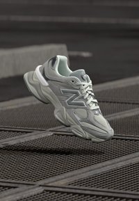 Ljust grå atletisk sko med en texturerad ovandel, mesh-detaljer och en kraftig sula. Har en framträdande logotyp på sidan och matchande snören.