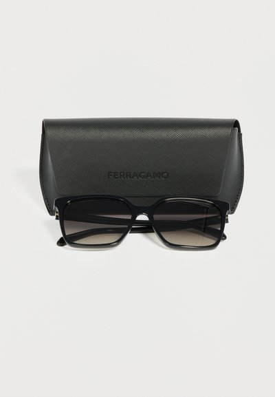 Lunettes de soleil rectangulaires noires avec verres teintés reposant devant un étui noir texturé de marque Ferragamo.
