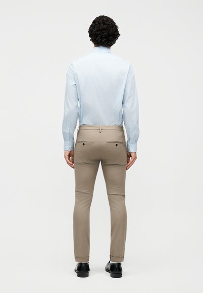 Άνδρας με σγουρά σκούρα μαλλιά που φορά ανοιχτό μπλε ριγέ μακρυμάνικο πουκάμισο, μπεζ παντελόνι slim-fit και μαύρα παπούτσια, στέκεται με την πλάτη.