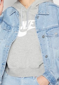 Chaqueta de mezclilla de color azul claro, con botones a presión y detalles desgastados, sobre una sudadera gris con un logo de Nike blanco. Texturas de tela suave.