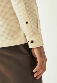 Manche de chemise beige texturée avec un motif en grille, dotée de boutons sombres aux poignets. Main visible positionnée près du côté.