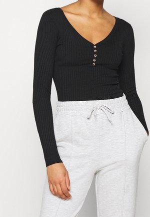 Mujer con una camiseta negra de manga larga acanalada con botones y pantalones deportivos gris claro con cordón, con la mano en el bolsillo.