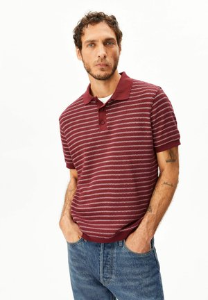 Mann mit lockigem Haar und leichtem Bart, der ein rot-weiß gestreiftes Poloshirt und blaue Jeans trägt und mit den Händen in den Taschen steht.