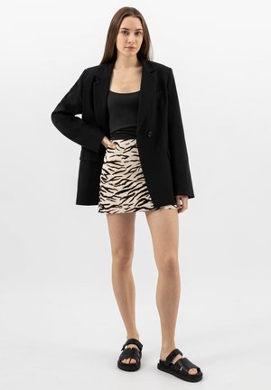 Jonge vrouw die een zwart colbert, een zwart topje, een minirok met dierenprint en zwarte sandalen draagt, staande tegen een effen witte achtergrond.