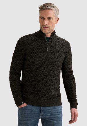 Hombre de mediana edad con cabello gris, vestido con un suéter oscuro de punto con cremallera hasta la mitad y pantalones vaqueros azules, de pie con una mano en el bolsillo.
