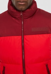 Primer plano de una persona que lleva una chaqueta acolchada roja y burdeos con un alto cuello y un parche del logo "HILFIGER" en el pecho.
