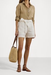 Light brown linen shirt with rolled sleeves, white linen shorts with a tied waist, brown strappy sandals, and a woven tote bag.