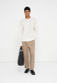 Krém színű kötött pulóver texturált mintákkal, bézs nadrág és fekete bőrtáska. A modell sima háttér előtt áll, a cipők díszített kialakítással rendelkeznek.